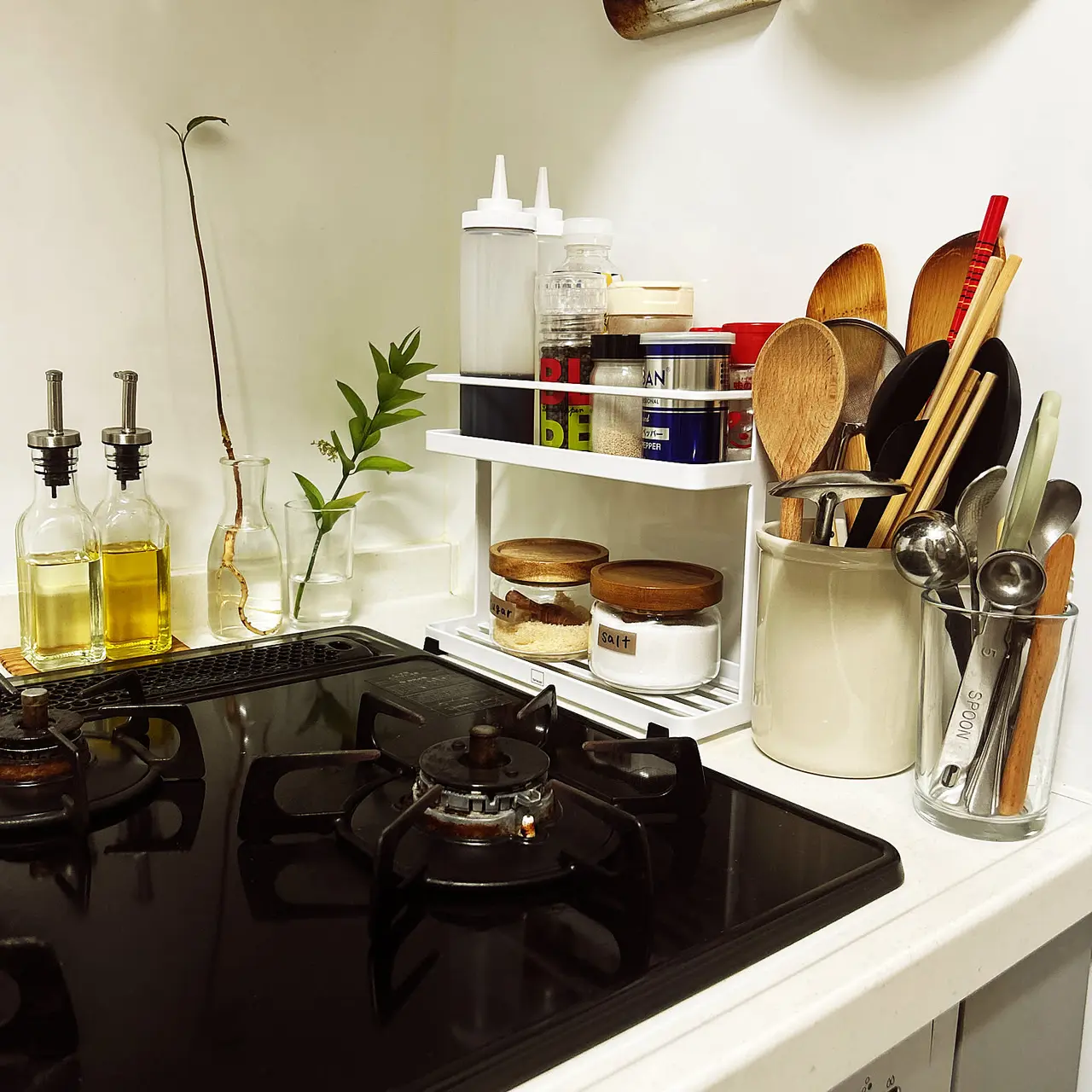 A white Yamazaki Tower spice rack beside a gas stove holding seasonings, jars, and small kitchen tools.