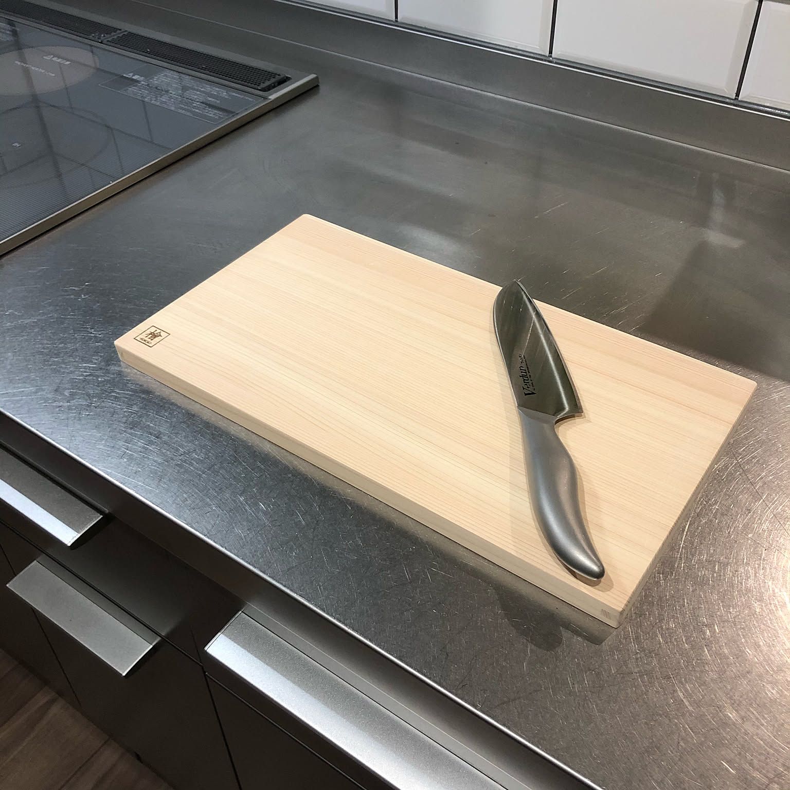 A pale rectangular hinoki cutting board with a Japanese kitchen knife resting on it, on a stainless steel kitchen counter.