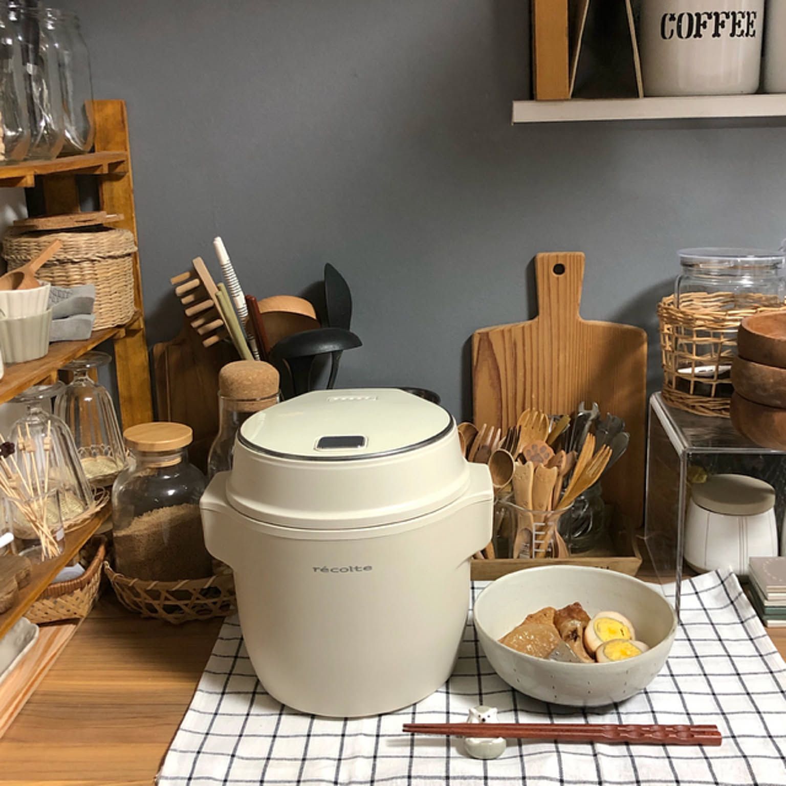 A compact white Recolte rice cooker on a kitchen counter with dishes and wooden utensils.