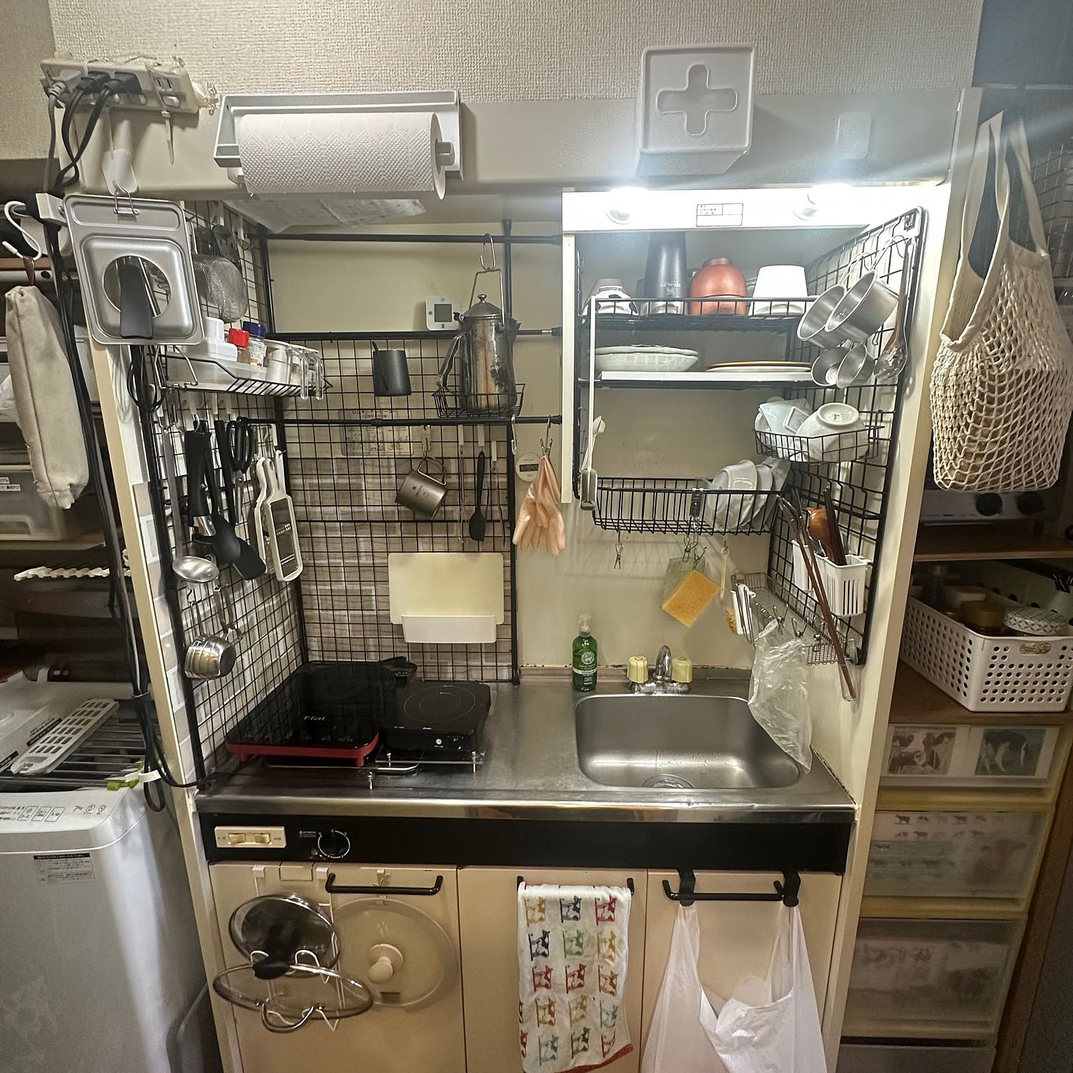 A compact Japanese apartment kitchen using wire panels, tension rods, baskets, and open shelves to organize tools, dishes, and cookware around a tiny sink and cooktop.