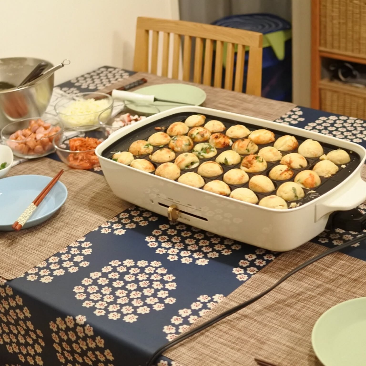 A BRUNO hot plate cooking takoyaki on a dining table with bowls of ingredients nearby.