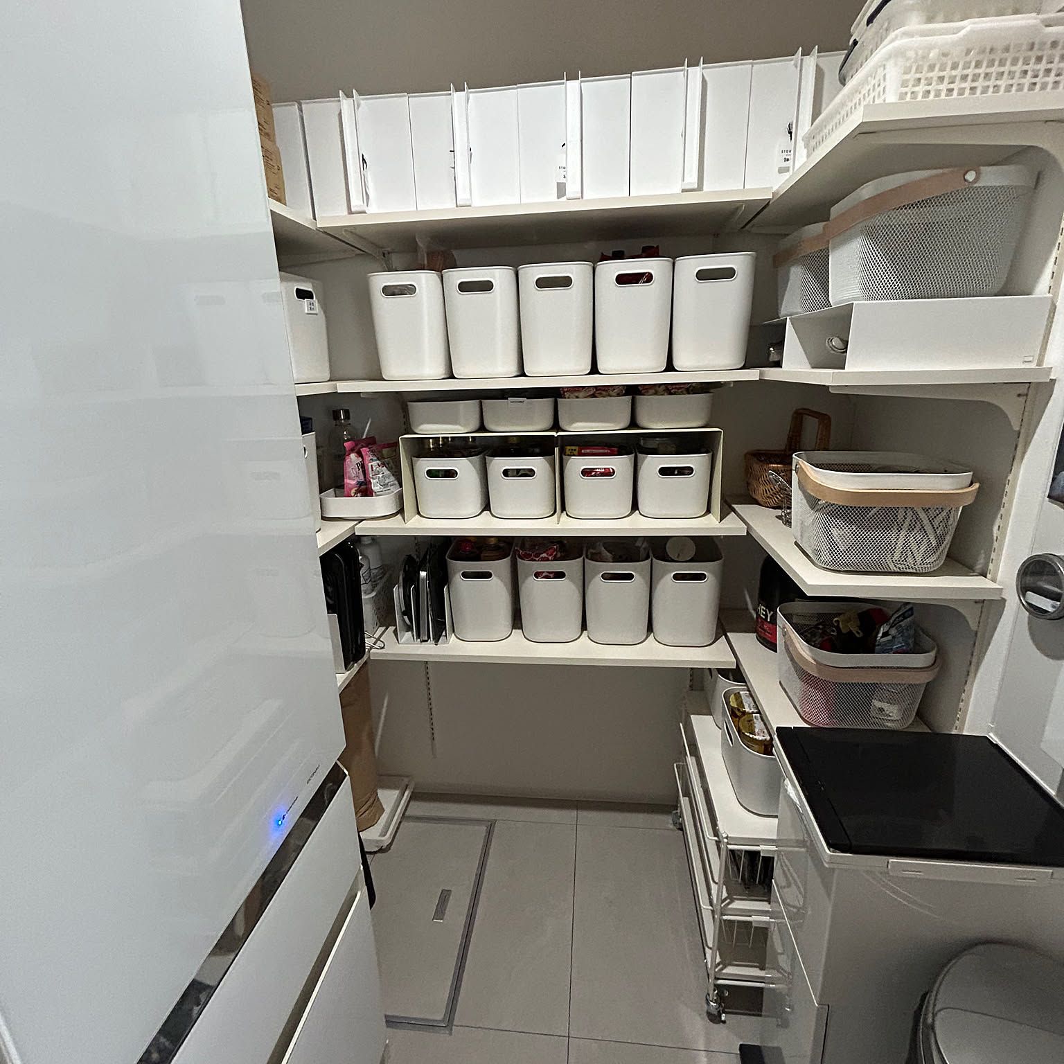A compact pantry shelf with white labeled bins, baskets, and food stock stored beside a kitchen