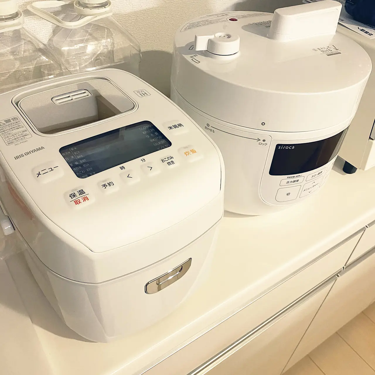 A white 3-cup rice cooker shown prominently beside another countertop cooker in a bright kitchen.