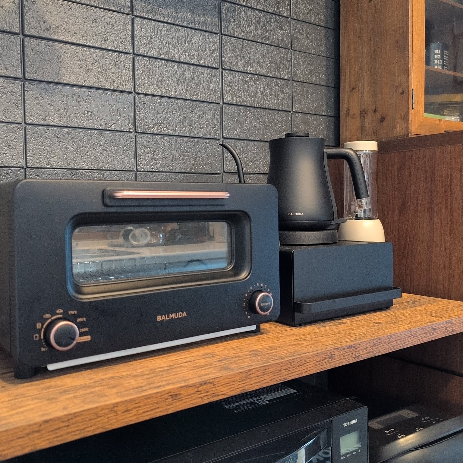 A black BALMUDA steam toaster and a black BALMUDA gooseneck kettle side by side on a wooden shelf in a Japanese kitchen