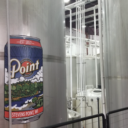 Large stainless steel brewing tanks stand inside a brewery facility, featuring a colorful “Point Brewery – Stevens Point, WI”