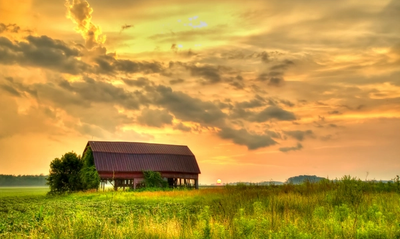 The sun sets over an old barn in a grassy field.