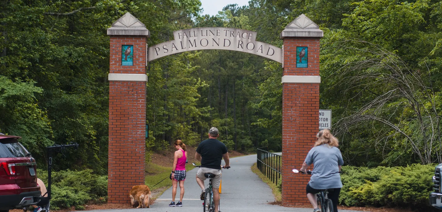 Cyclists and a dog walker enjoy the scenic, tree-lined trail at the Psalmond Road entrance to Fall Line Trace, a popular path for biking and walking in Columbus, Georgia.
