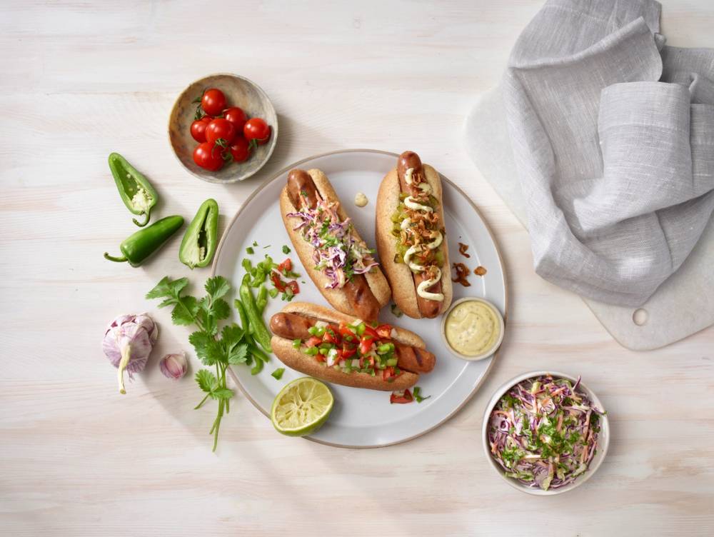 Tre grillede pølser i brød med ulike typer tilbehør, som coleslaw, syltetøy og sauser, presentert på et lyst bord.