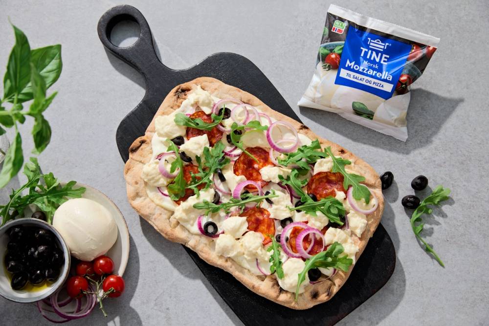 Pinsa Romana med mozzarella, pepperoni, rødløk, oliven og rucola på et svart skjærebrett.
