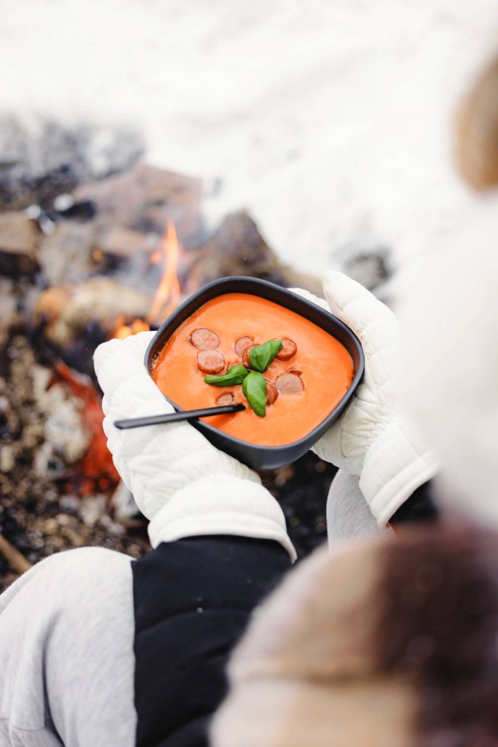 Hender med votter som holder en bolle tomatsuppe med grillepølser ved et bål om vinteren.