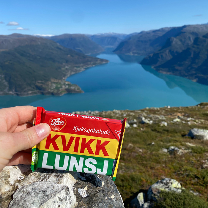 Hånd som holder Kvikk Lunsj tursjokolade i fjordlandskap.