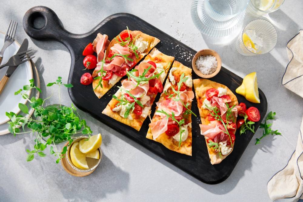 Pinsa Romana skiver med prosciutto, tomater og rucola på et svart skjærebrett.
