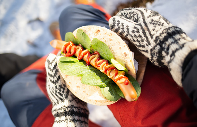 Hender med ullvotter som holder pølse i lompe med ketchup og sennep.
