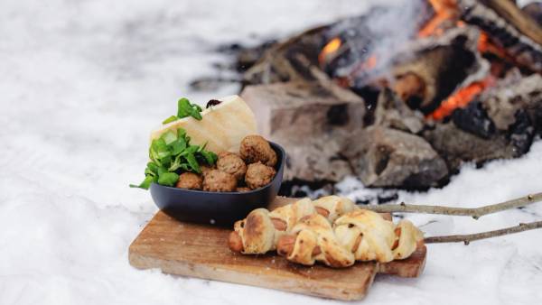 Pølse med pinnebrød og kjøttboller med pitabrød foran bål i skogen om vinteren.