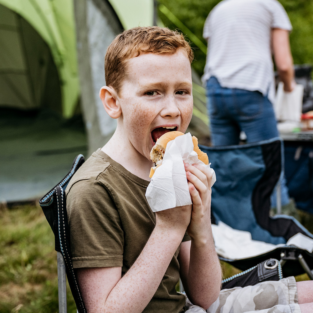 Gutt som spiser burger foran telt.