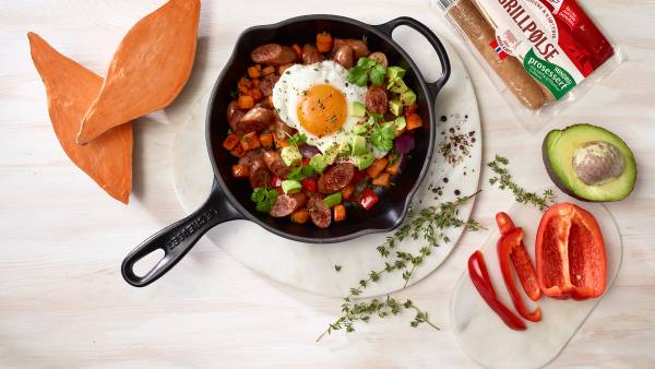 En stekepanne med Gildes grovere grillpølser, søtpotet, paprika, avokado og stekt egg, omgitt av ferske ingredienser på et lyst bord.