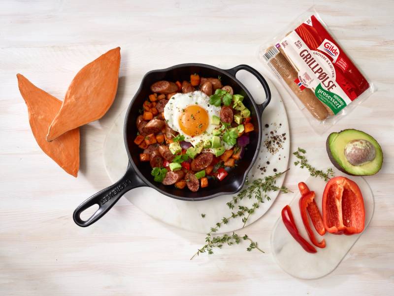 En stekepanne med Gildes grovere grillpølser, søtpotet, paprika, avokado og stekt egg, omgitt av ferske ingredienser på et lyst bord.