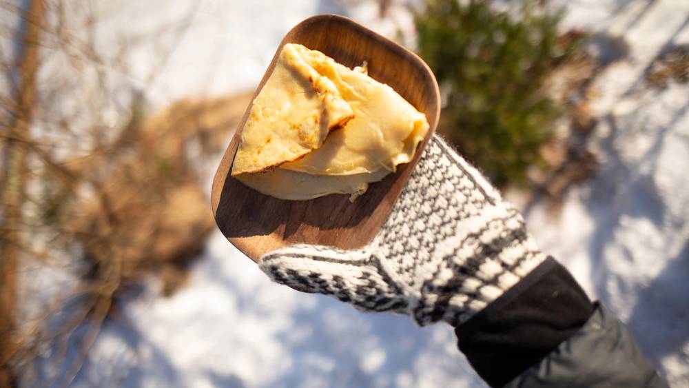 Hånd med vott som holder en tallerken med pannekake på tur i skogen.