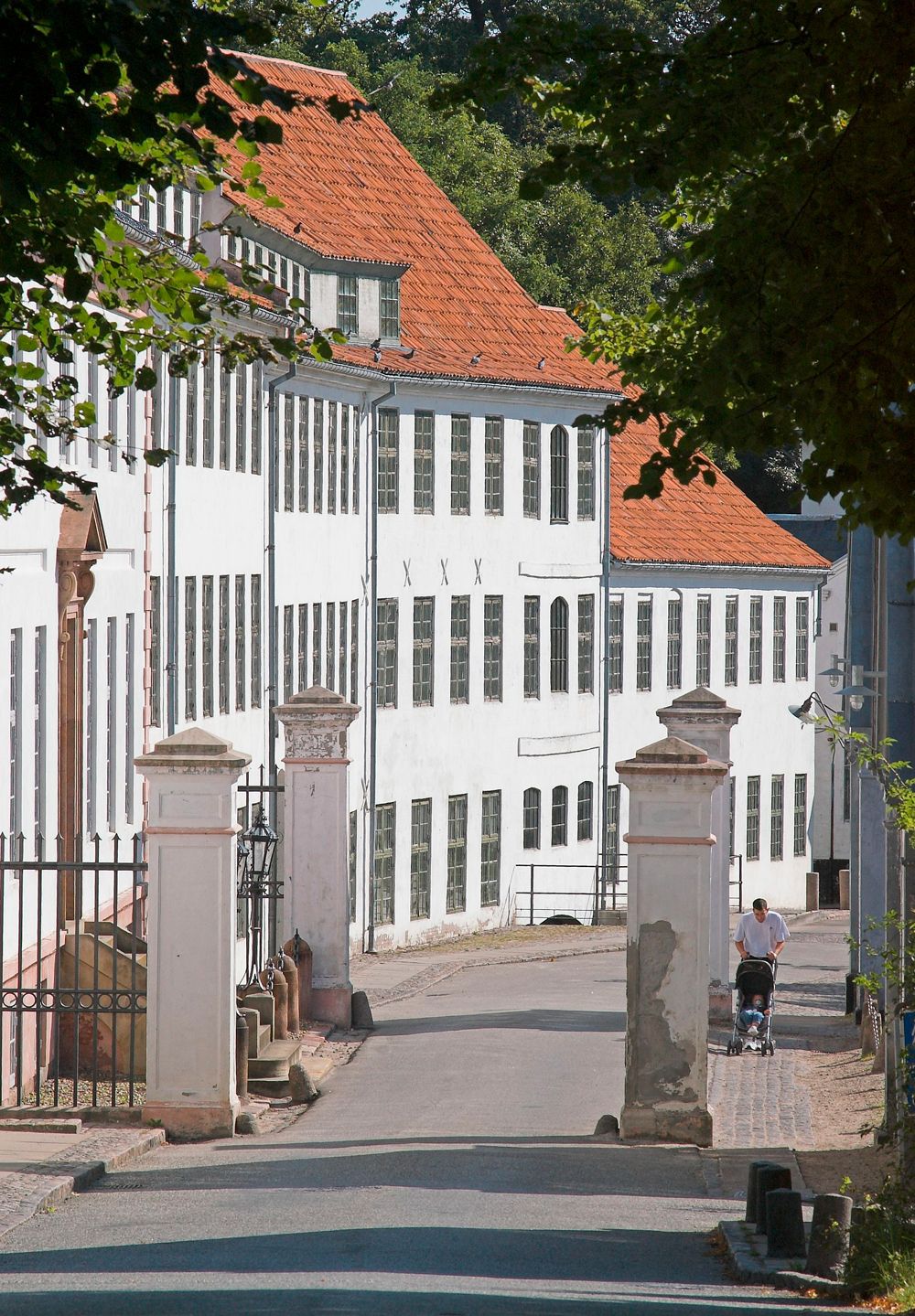 Hvid fabriksbygning med rødt tegltag ved indkørslen til Brede Værk set fra vejen gennem den gamle portåbning