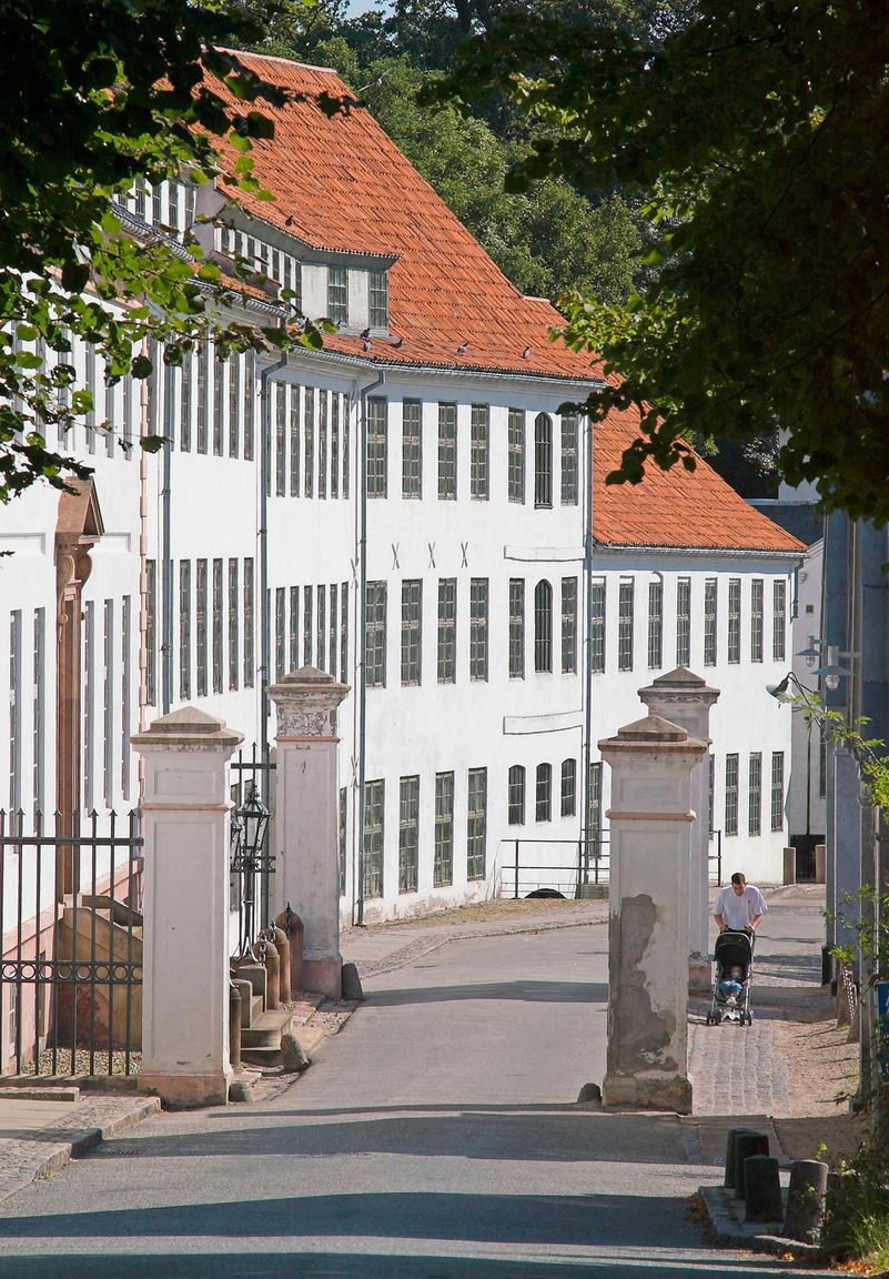 Hvid fabriksbygning med rødt tegltag ved indkørslen til Brede Værk set fra vejen gennem den gamle portåbning