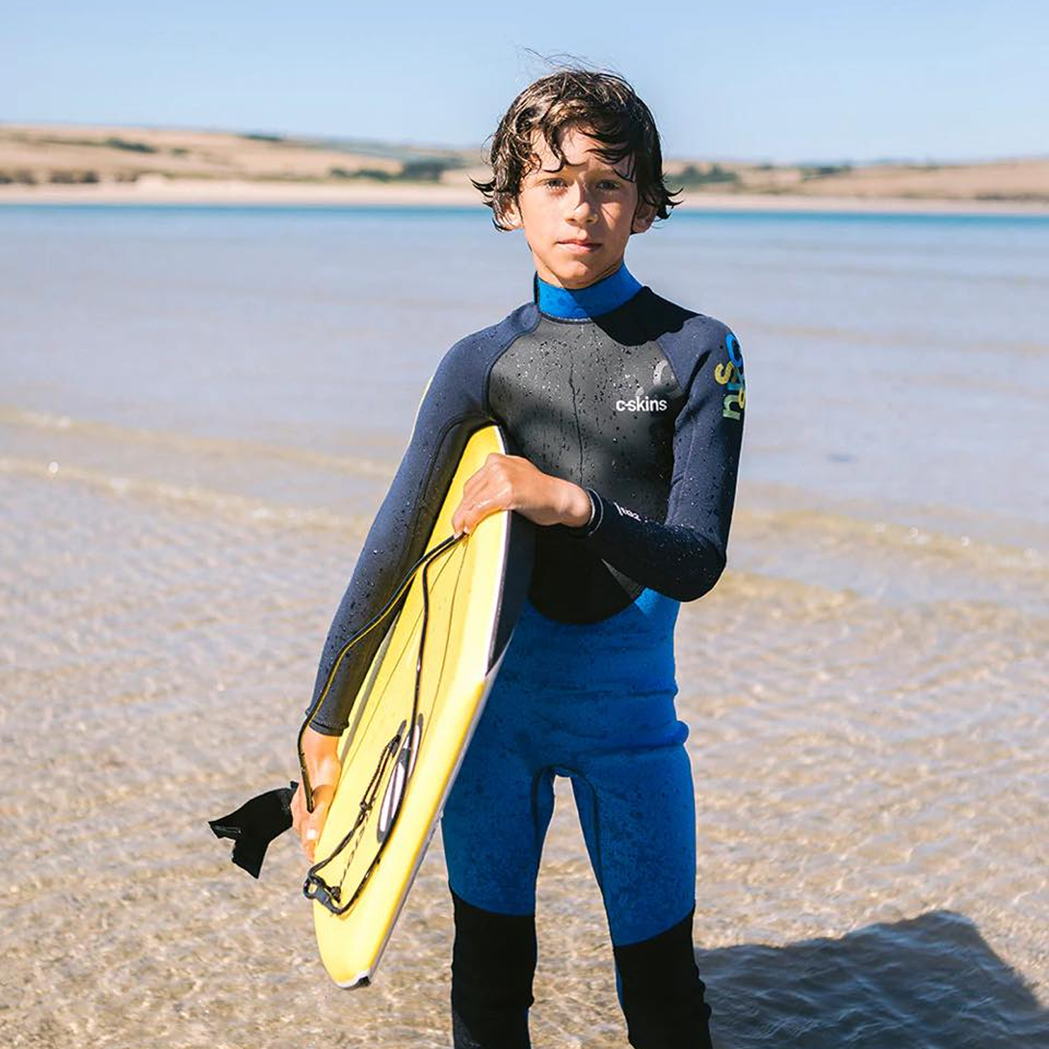 Young kid on beach holding surfboard in blue and black wetsuit