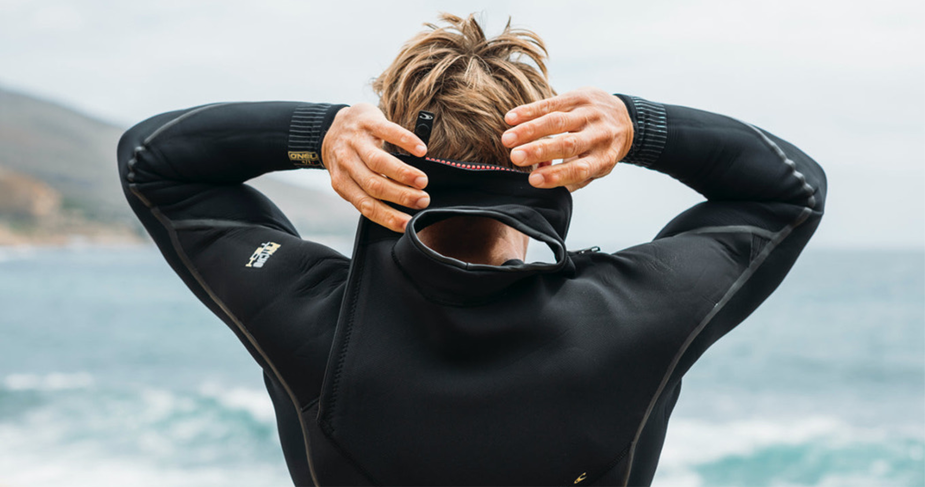 Man on beach in wet suit from behind with hands behind head