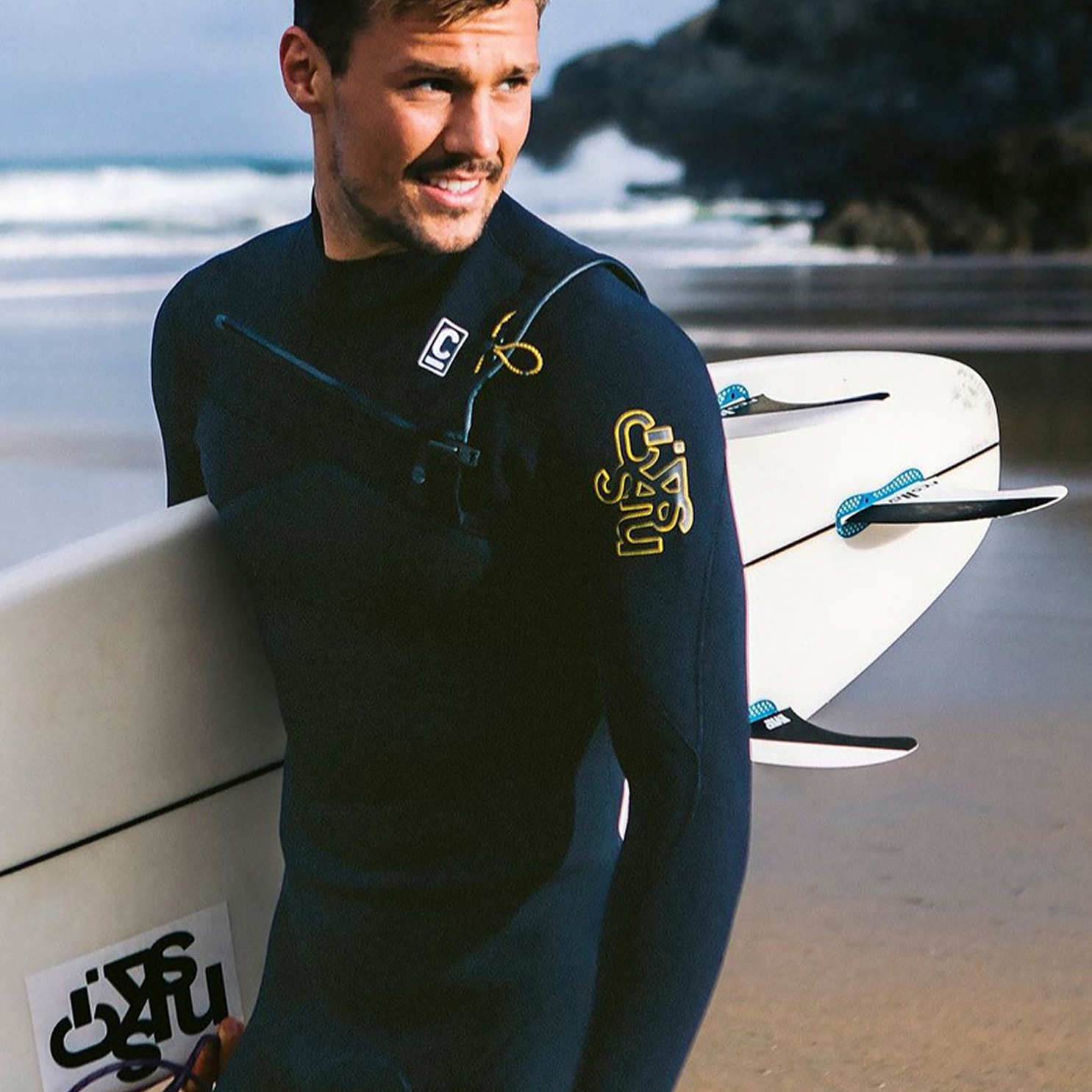 Man holding surfboard smiling on beach in back wetsuit with yellow logo on arm
