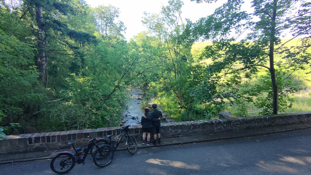 The first bridge over the Water of May Two cyclists stand looking over the edge of an old stone bridge