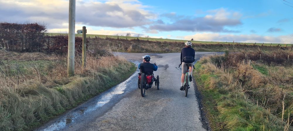 a photo of an adaptive cyclist and two wheel cyclist turning from a small road onto a farm track