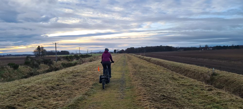 a photo of a cyclist towing a trailer on an old disused railway with flat views of the countryside either side.