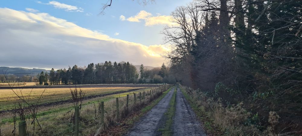 photo of a gravel track with woods on one side and fields on the other with views of the hills in the distance