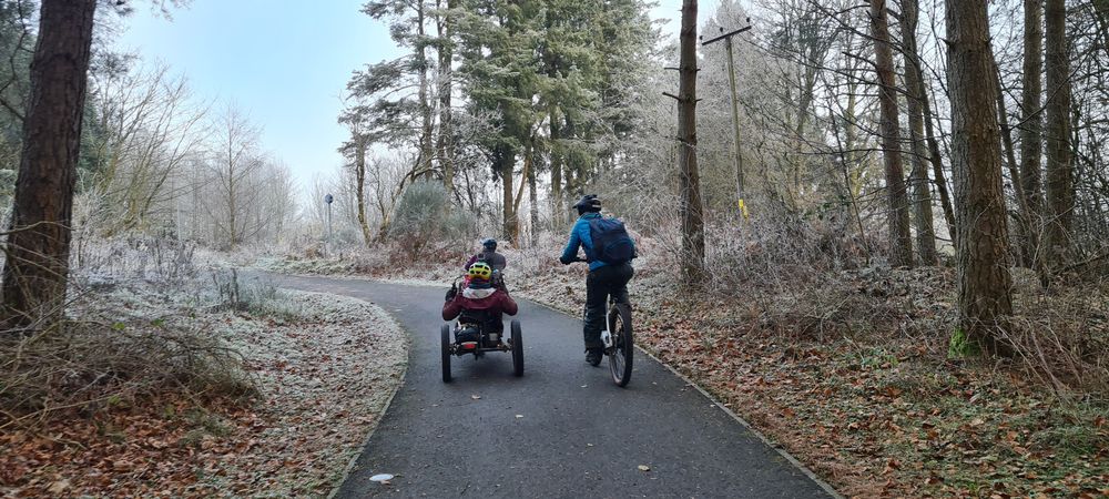 photo of two adaptive riders and a two wheel cyclists on a tarmac multi-use trail in the trees