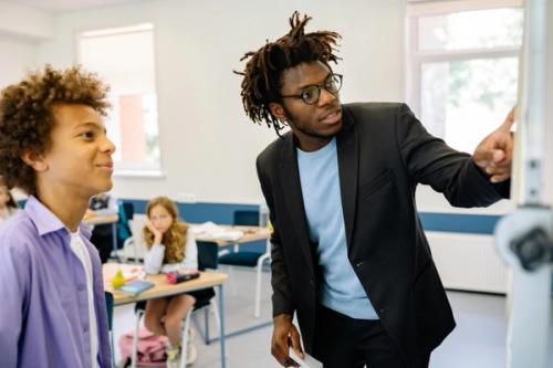 A man standing by a white board at the front of a classroom pointing to something on the board. A child stands next to the man, smiling at what they are looking at.
