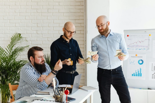 Three men with glasses standing in an office holding money and talking. There are charts in the background with figures of how much earnings are being made.