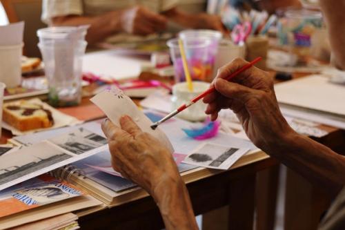 A pair of hands holding a paper and using a watercolor paintbrush to paint.