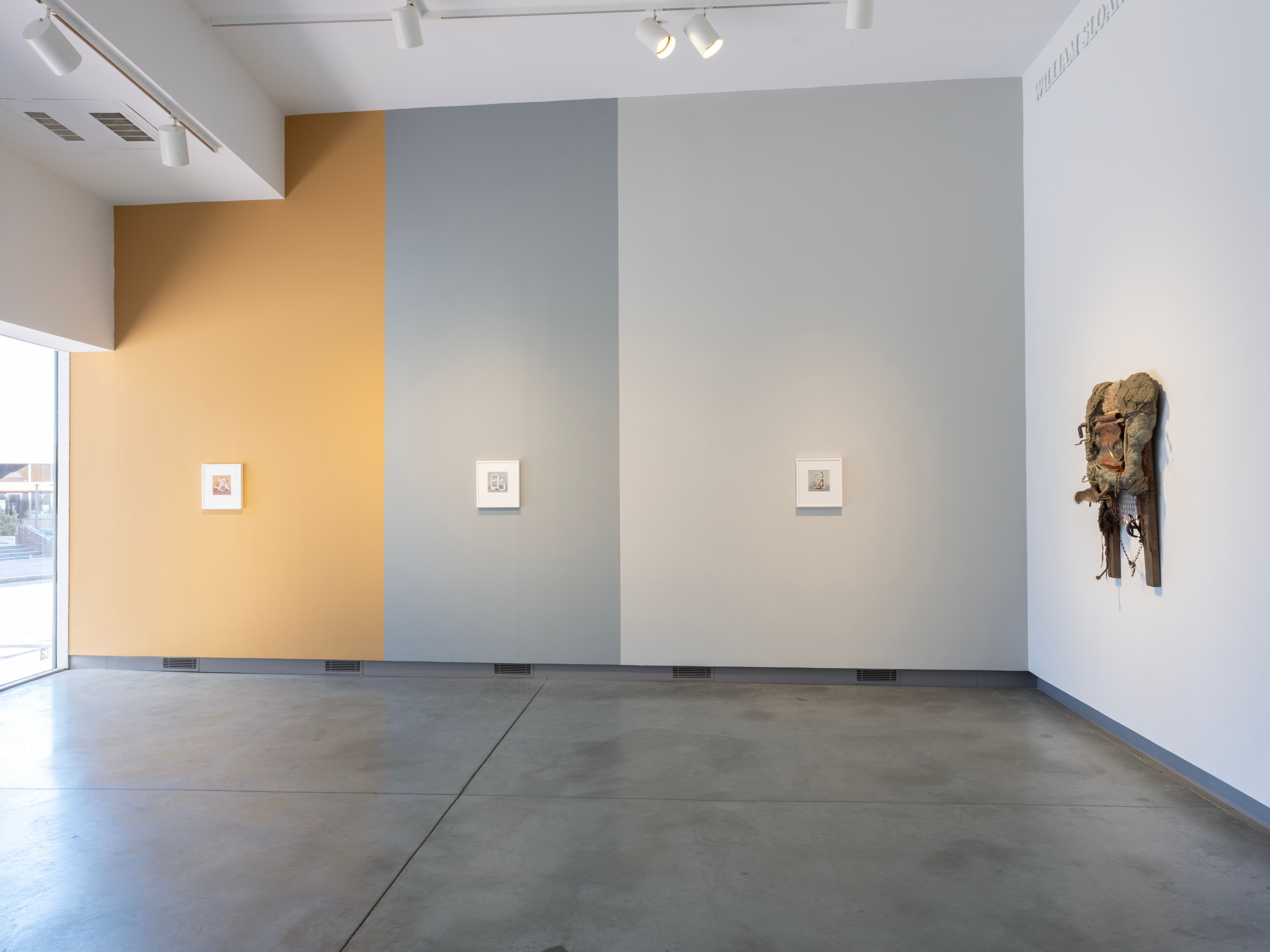 ENTER: installation view of three framed photographs against a wall painted yellow, dark grey, and light grey, and a sculpture hanging from the wall.