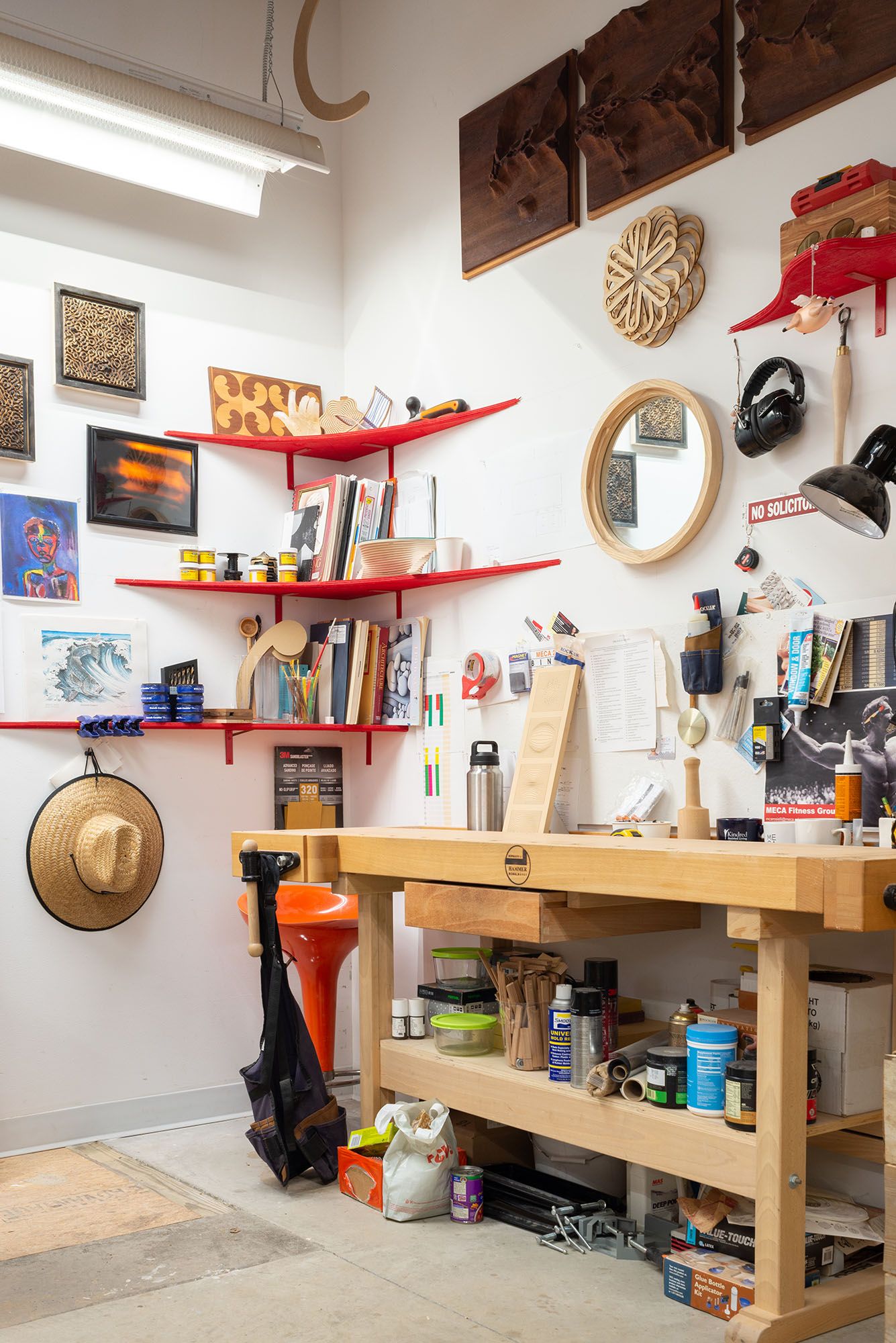 A woodworking studio space with a workbench, wooden sculptural pieces on the wall, and floating red shelves holding books.