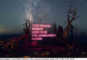 A neon pink sign with the words, "This present moment used to be the unimaginable future" against a starry night sky.