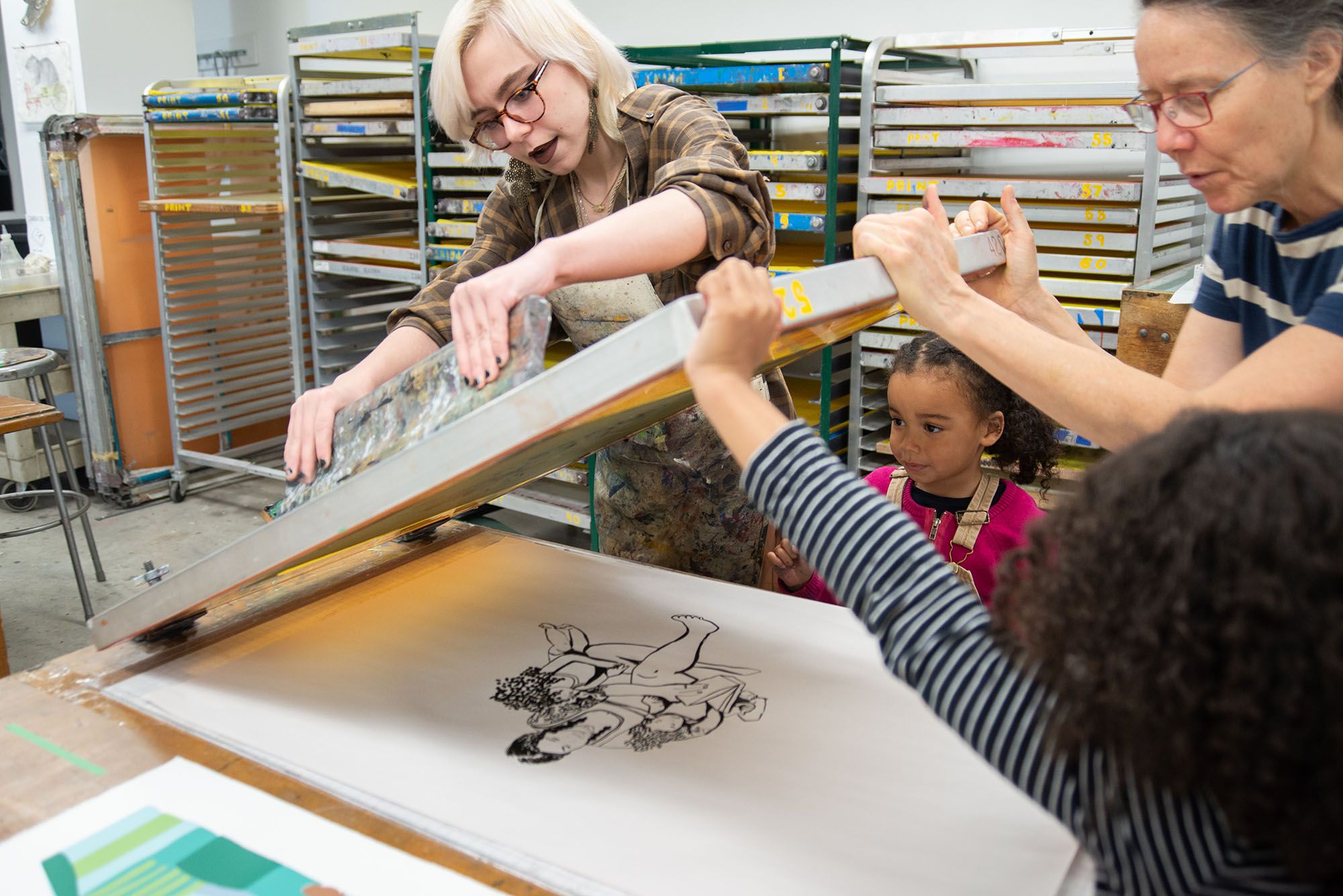 A student, professor, and two young children lifting a screen off of a screenprint depicting a mother with her two daughters.