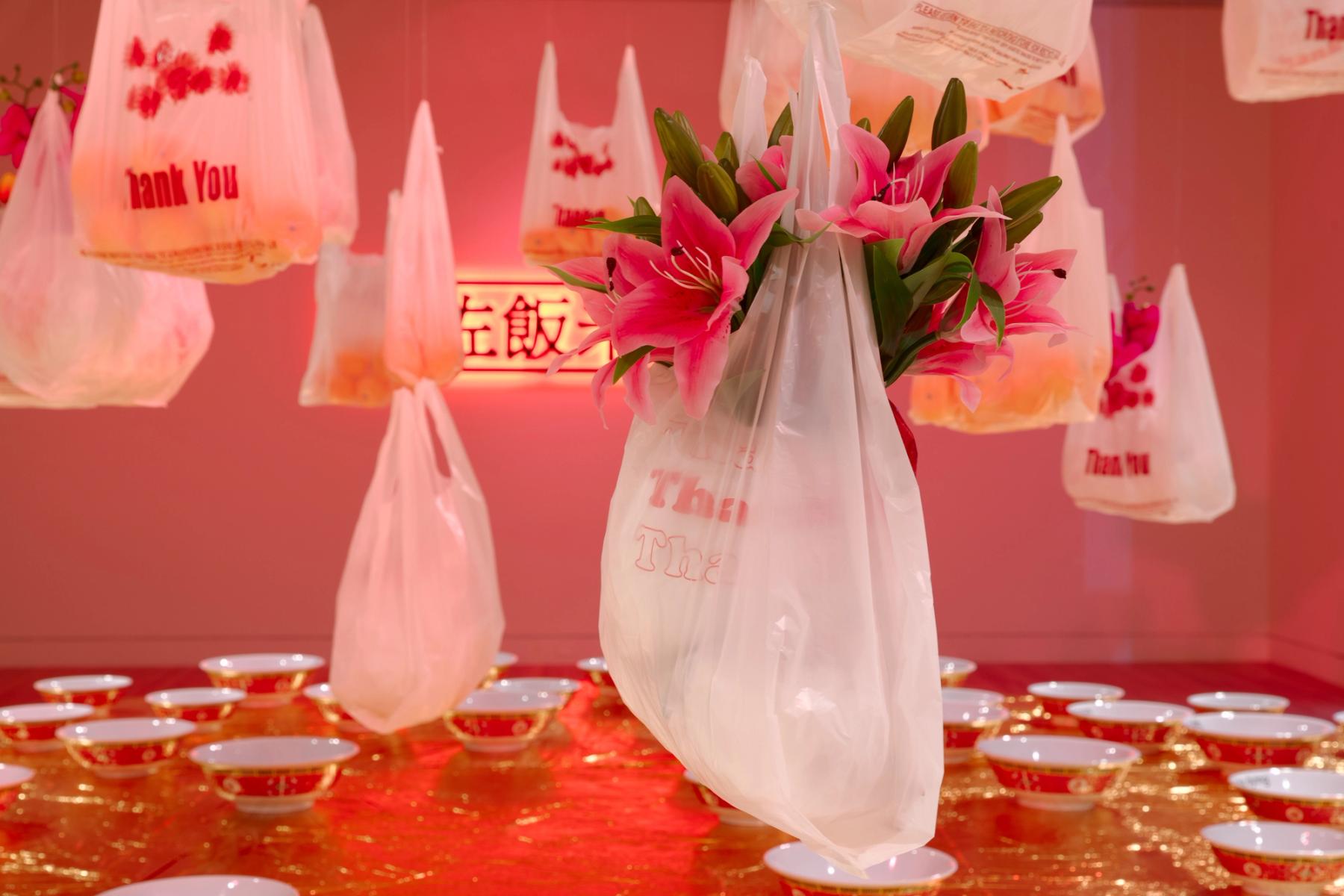 An installation of hanging plastic grocery bags filled with pink lilies over a table of red bowls.