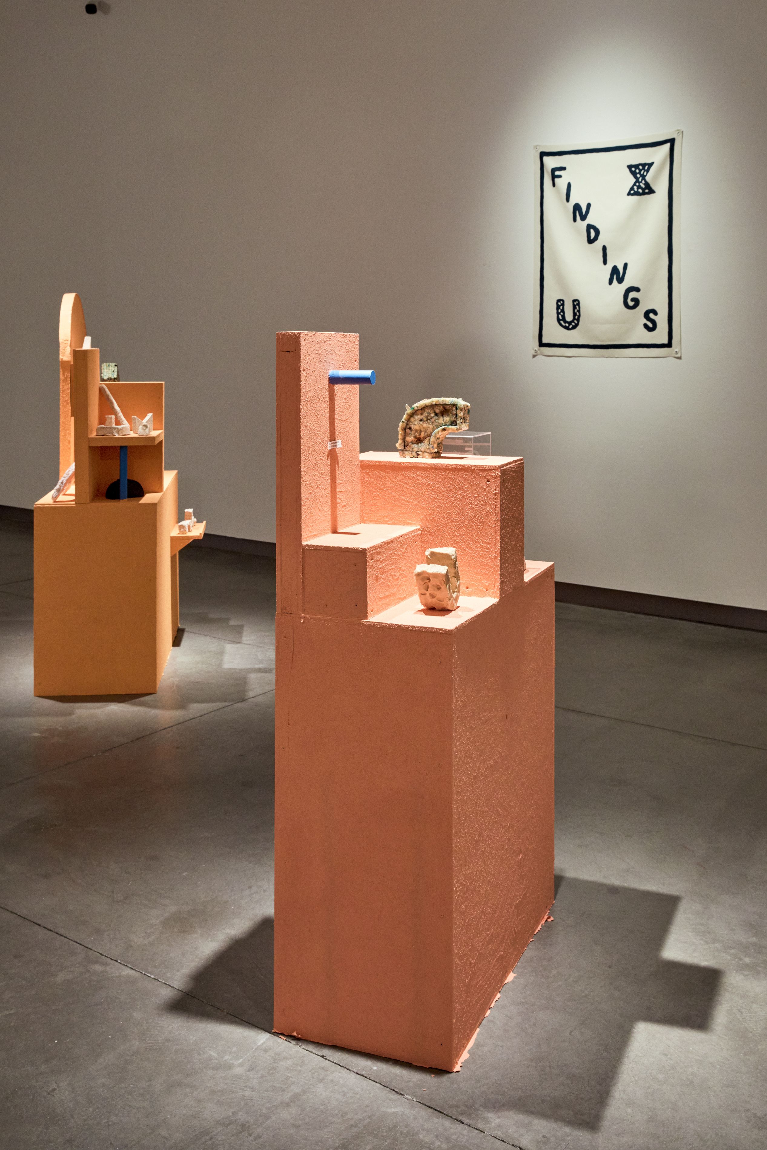 Gallery view of two orange sculptures and a wall hanging with the word, "Findings"