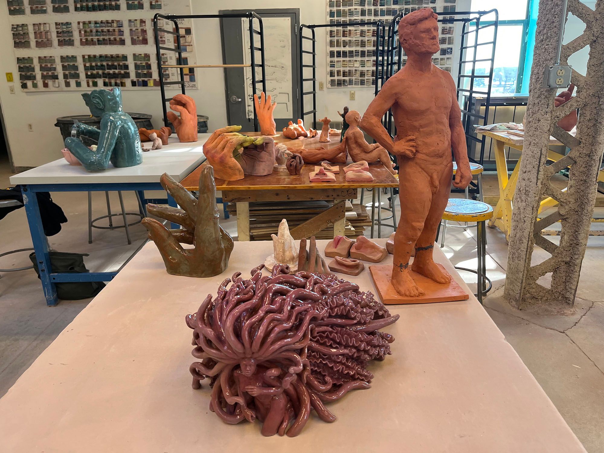 View of in-process ceramic sculptures including a medusa-like figure, a nude man, several hands, and a dispersed face.