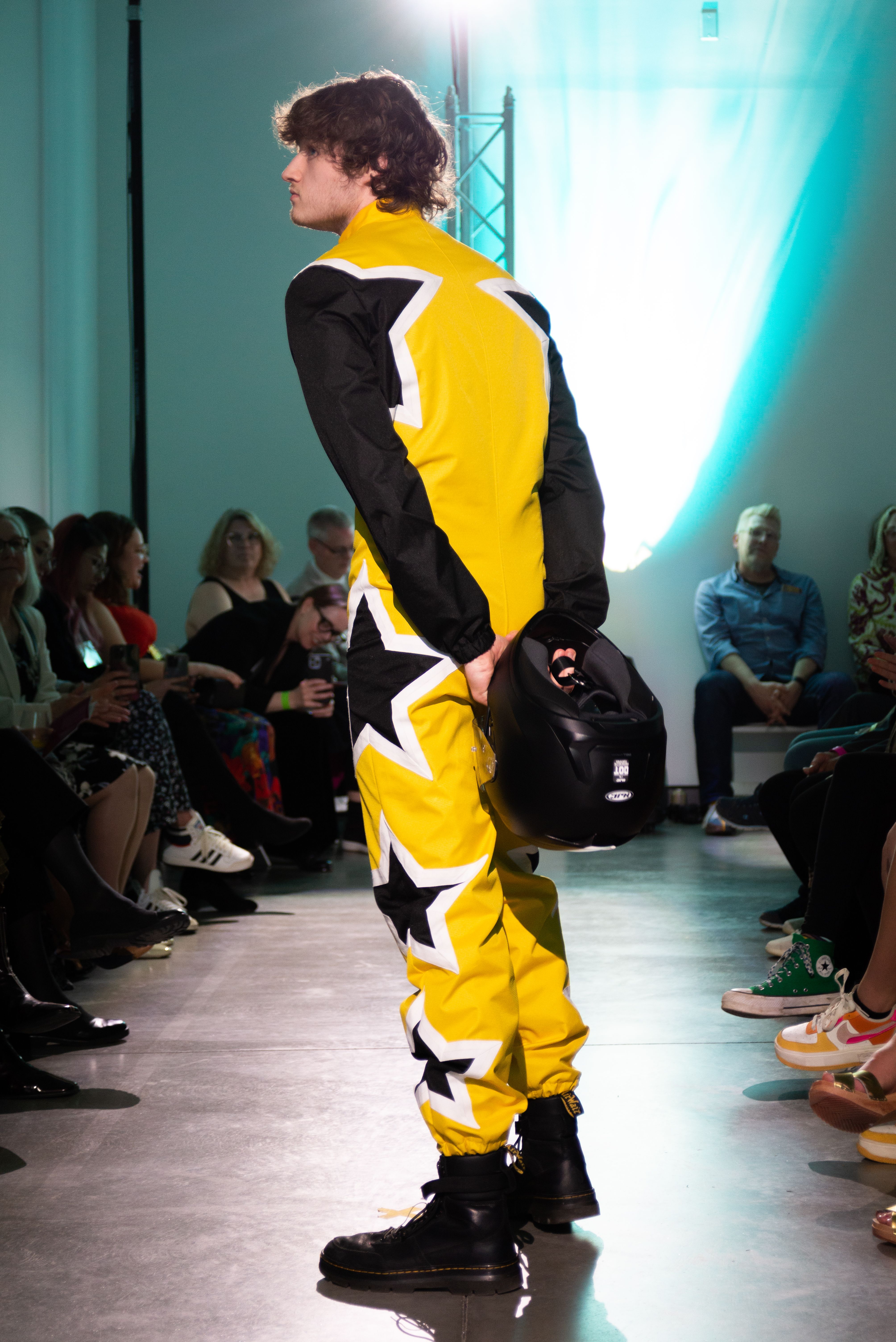 A model on the runway wearing a yellow jumpsuit with black stars down the legs, holding a motorcycle helmet.