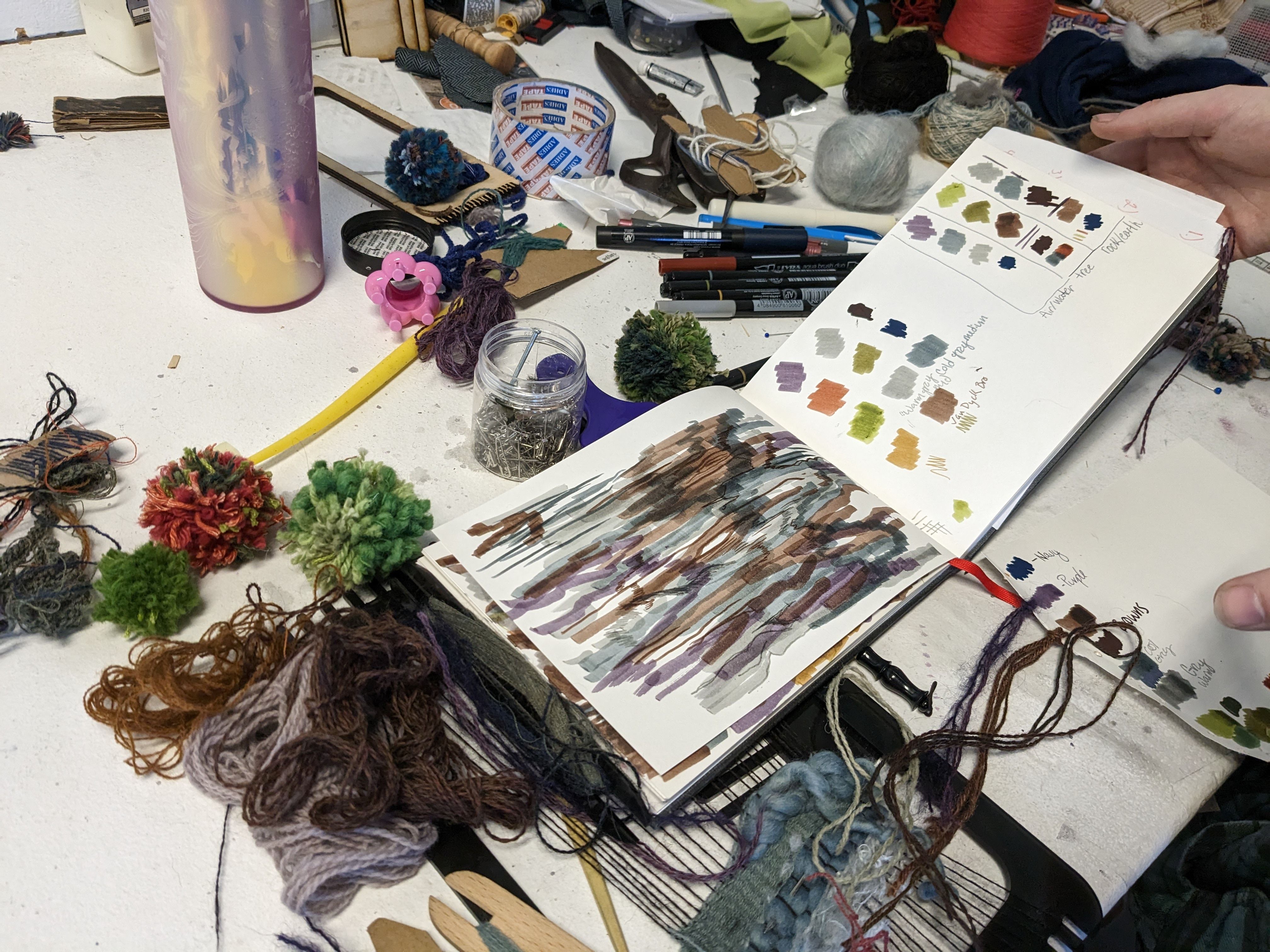 An open sketchbook with color swatches on a desk covered with different colors of yarns and pompoms