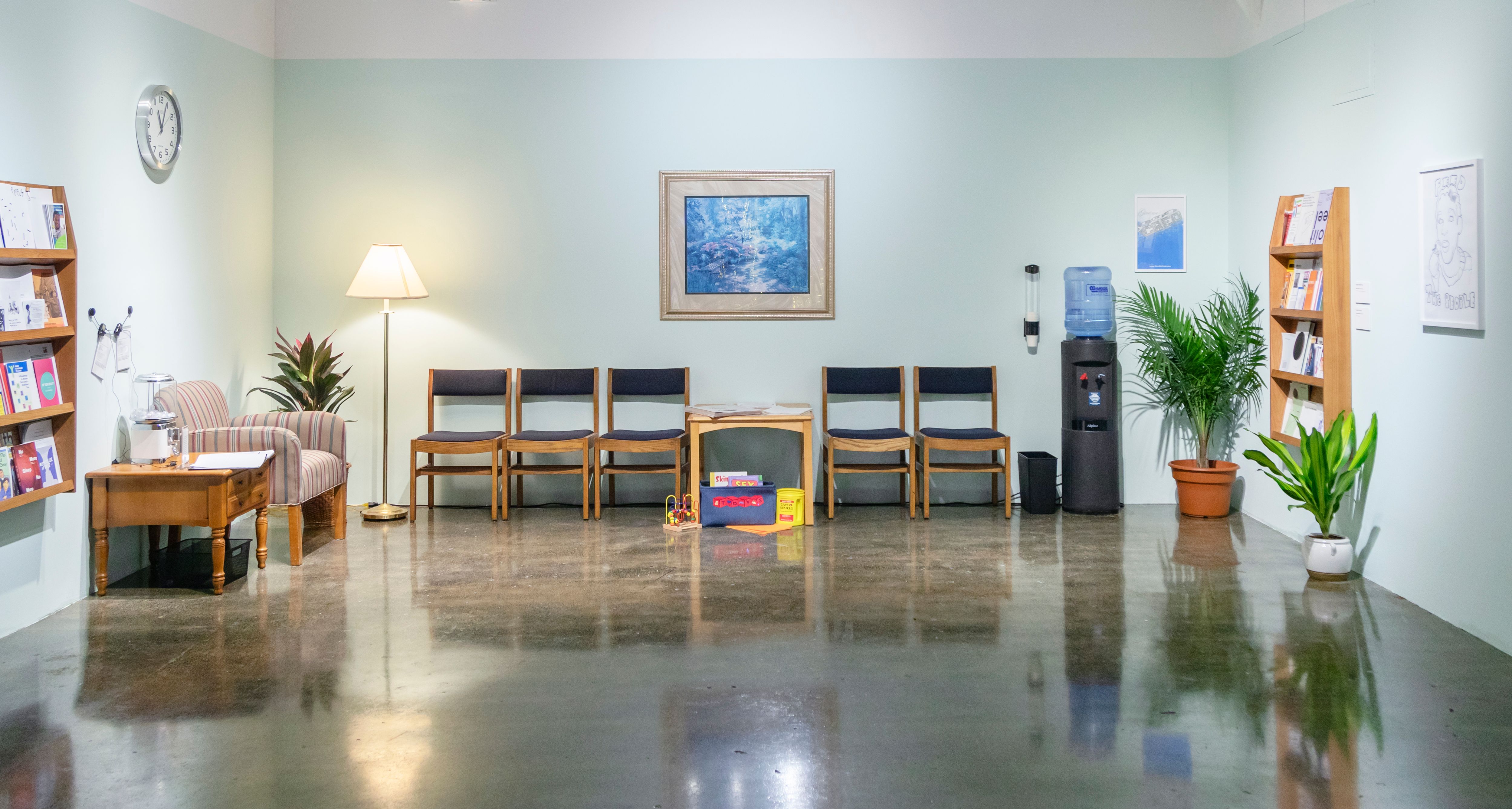 A photograph of The Waiting Room in “Sick Time, Sleepy Time, Crip Time,” curated by Taraneh Fazeli. The image includes bookshelves, wooden chairs, children's toys, a water fountain, plants, a clock, art on the walls, and a floor lamp. The floor is a shiny, dark grey and the walls are a light blue-green. All furniture is on the edges of the room, with the center left empty.