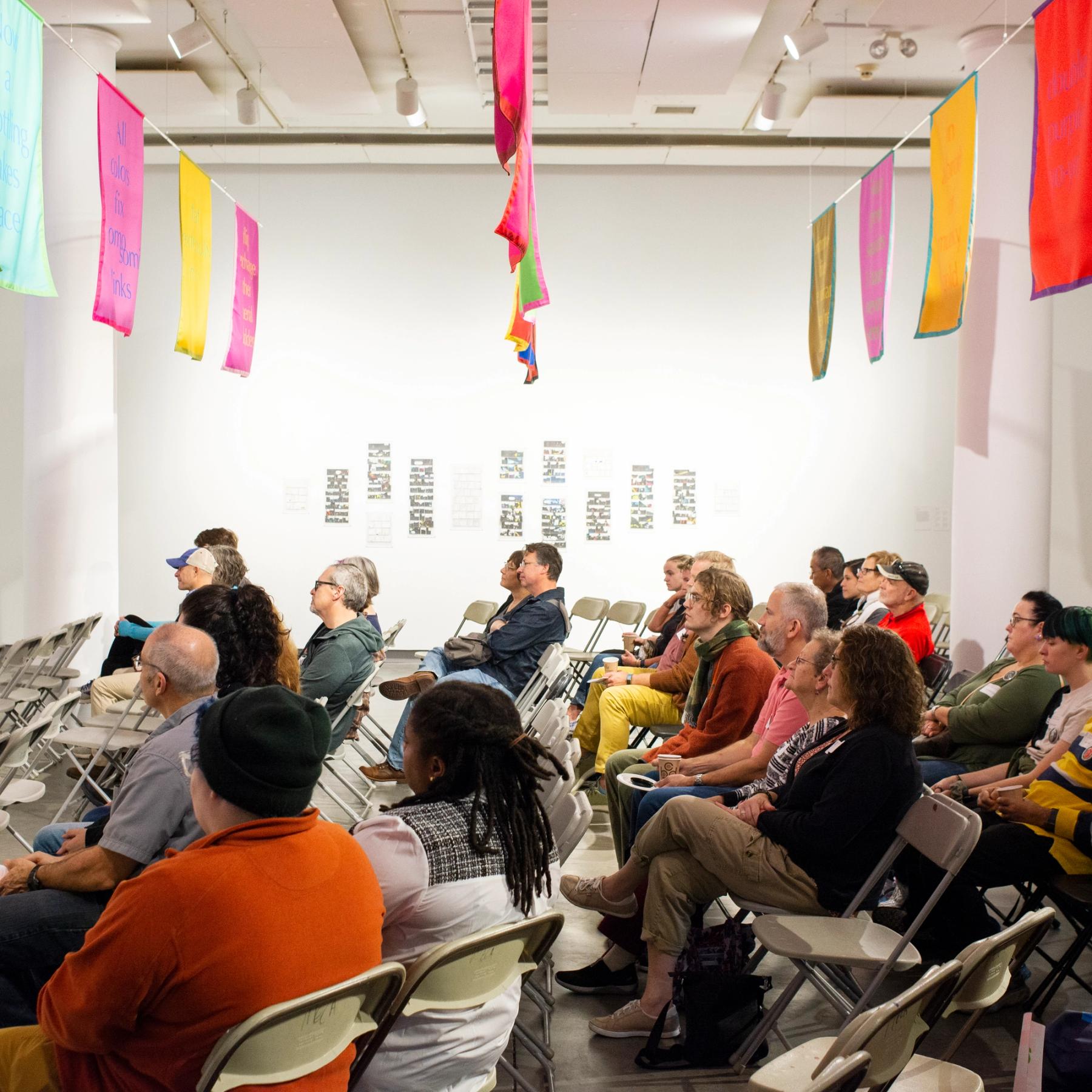 A croud of people sitting in chairs, listening, at the ICA with colorful flags above them.