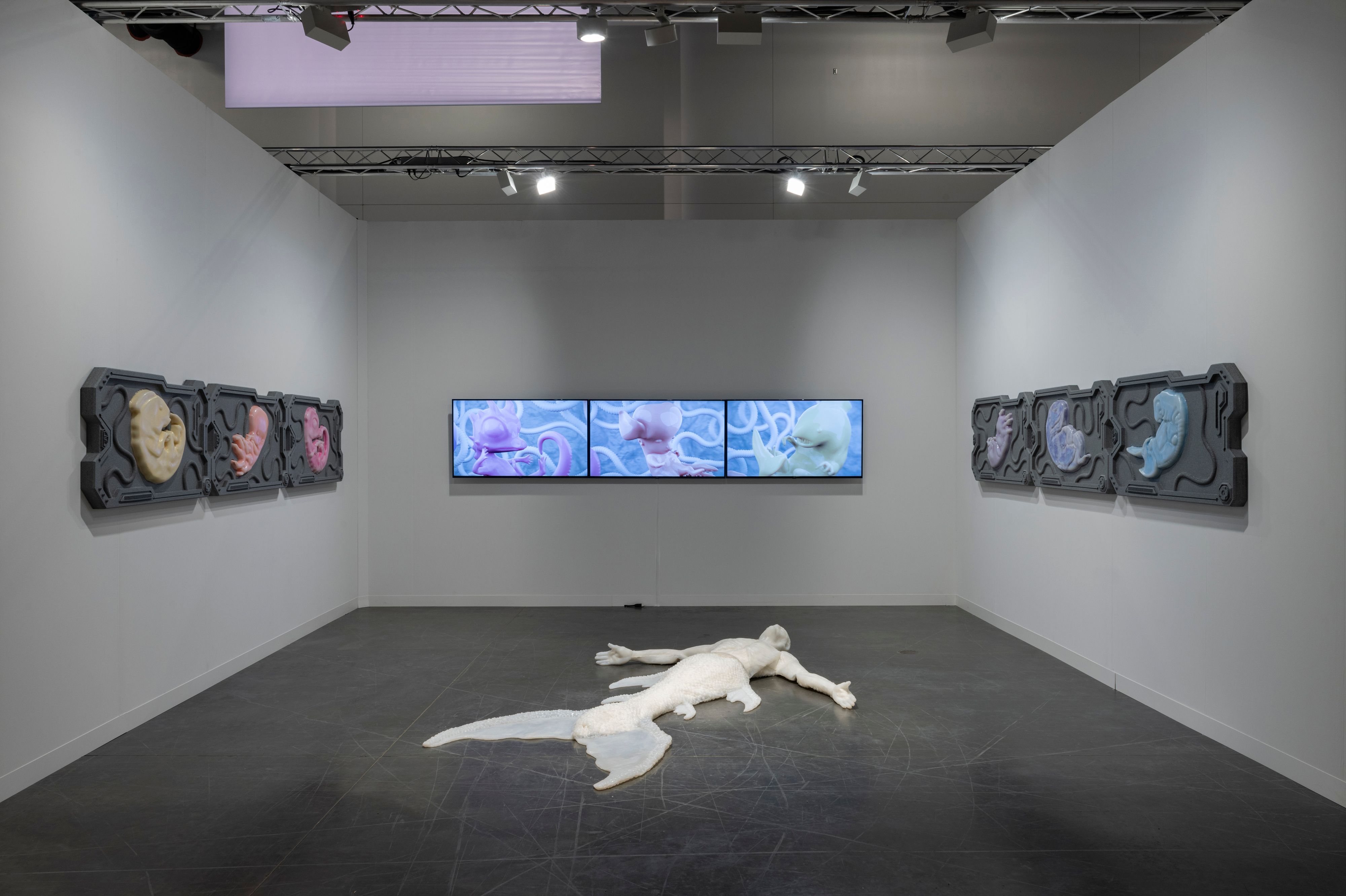 An installation view of a life-sized white silicon mermaid laying on the floor surrounded by screens of fetal figures.