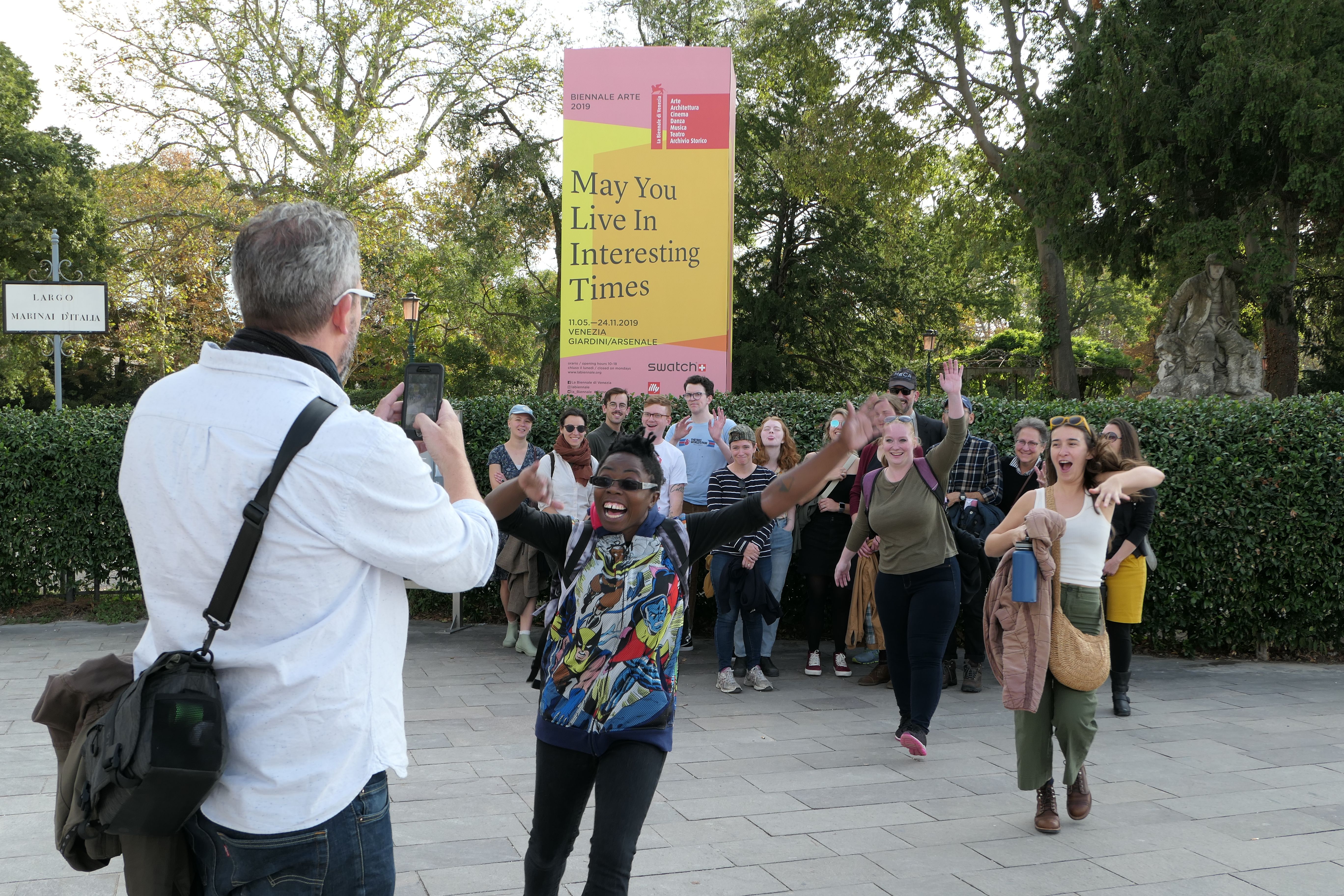 A group of excited MFA students in front of a sign that reads "May You Live in Interesting Times"