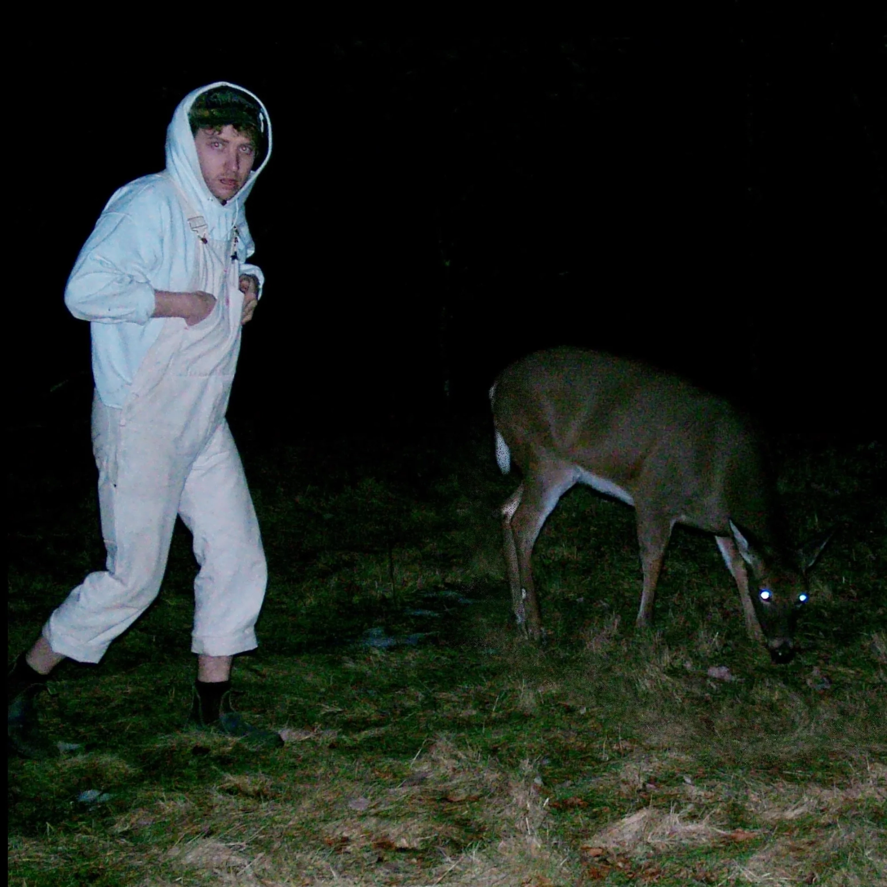 A photograph of a person in a white hoodie and white overalls next to a deer eating grass with shining eyes at night.
