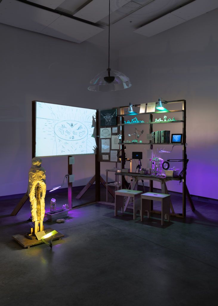 Installation view of shelves with scientific books and objects next to a whiteboard projection and a sculptural figure.